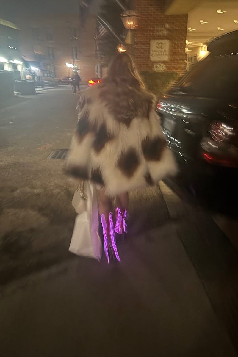 A person walking on a city street at night, viewed from behind, wearing Cowboy LED boots, a large brown-and-white patterned faux-fur coat and carrying a white tote bag. A black car is parked on the right, and the street and hotel exterior are visible in the background.