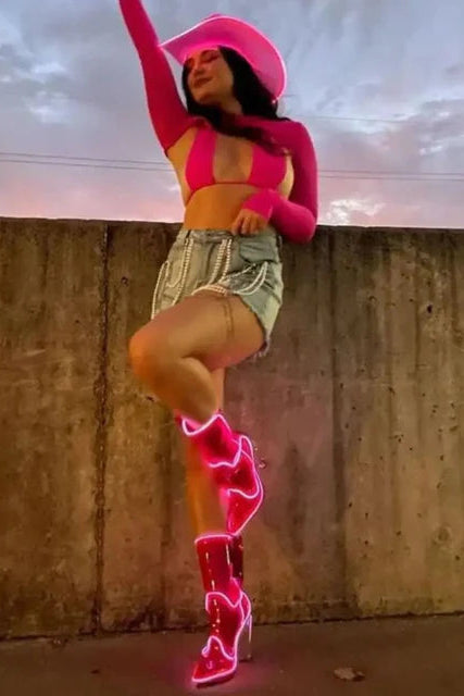 A woman posing outdoors against a concrete wall, wearing a bright pink cowboy hat, pink long-sleeve crop top, denim shorts, and pink high-heeled Cowboy LED boots with glowing neon-style pink outlines.