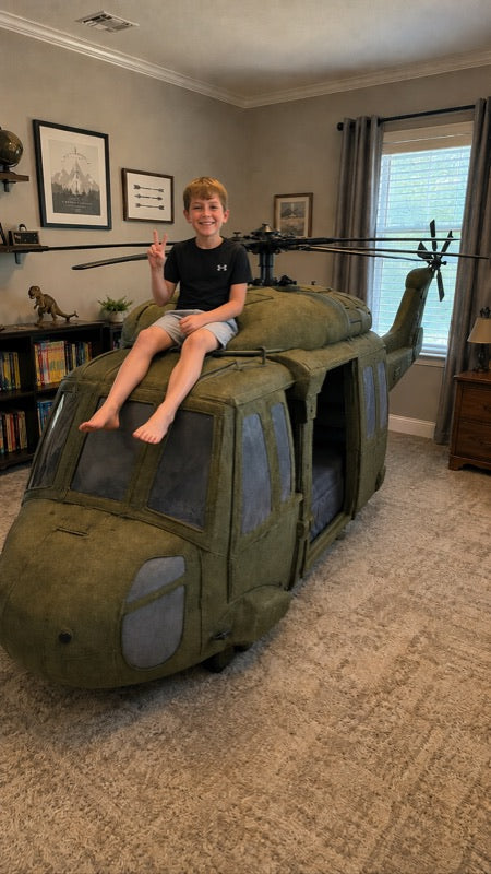 A young boy sits on top of a large olive green helicopter-shaped bed in a carpeted room with bookshelves and framed pictures on the wall.