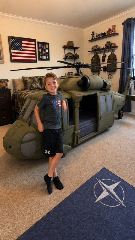 A young boy stands smiling in a child's bedroom next to a large olive green military helicopter bed, with a blue Navy-themed rug in the foreground.